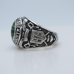May include: A silver-colored ring with a green gemstone. The ring features engraved text and symbols, including the words "Courage," "Honor," and "Conquer." The ring has a shield-shaped design with the letters "AF" in the center.