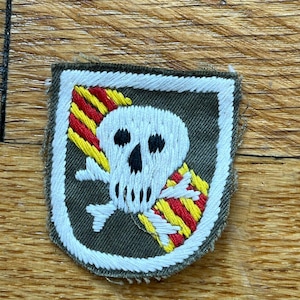 Può includere: Una toppa ricamata verde, bianca e rossa con un design di teschio e ossa incrociate. Il teschio è bianco con occhi neri e una bocca nera. Le ossa incrociate sono bianche con strisce rosse e gialle.