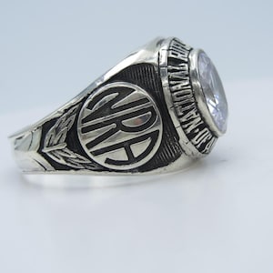 May include: Silver ring with a large, oval, light-colored stone. The ring features the letters "NRA" within a circle, surrounded by text that reads "NATIONAL RIFLE ASSOCIATION". A leaf design adorns the side of the ring.