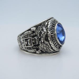 May include: Silver-toned ring with a large, oval blue gemstone. The ring features detailed engravings, including the words "United Naval Academy" and other text, along with an eagle and shield design. A classic piece of jewelry.