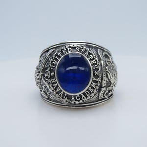 Op de afbeelding: Zilveren ring met een grote, ovale, diepblauwe steen in het midden. De ring is gegraveerd met de woorden "UNITED STATES NAVAL ACADEMY" en heeft gedetailleerde zijontwerpen, waarschijnlijk marinesymbolen.