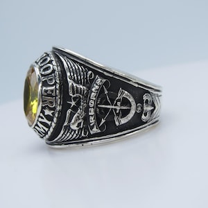 May include: A silver-colored ring with a large, oval, yellow gemstone. The ring features detailed engravings, including the words "MY ARMY" and "AIRBORNE," along with military symbols. The design suggests a military or commemorative theme.
