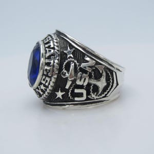 Könnte beinhalten: Ein silberner Ring mit einem großen, ovalen, blauen Edelstein. Der Ring zeigt die Worte "UNITED STATES NAVY" und ein Ankerdesign. Der Ring ist mit Sternen und einem strukturierten Band versehen.