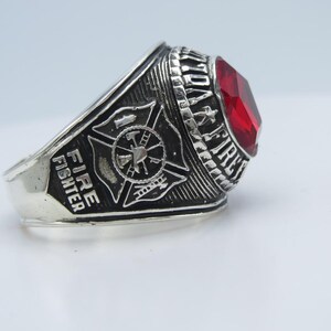 May include: Silver-toned Fire Fighter ring with a large, faceted red gemstone. The ring features the words "FIRE FIGHTER" and "TO PROTECT & SERVE" in raised lettering, along with a Maltese cross and fire tools emblem.