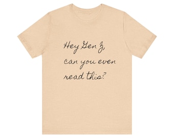 Puede incluir: Una camiseta de color beige claro con el texto "Hey Gen Z, can you even read this?" impreso en negro.