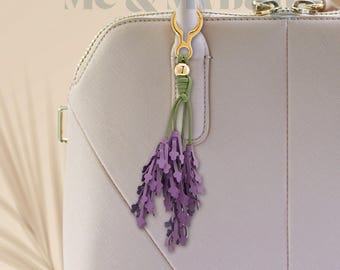 Puede incluir: Un charm de bolso de cuero color lavanda con forma de ramitas de lavanda, sujeto a un cierre dorado y un cordón verde. El charm cuelga de un bolso de mano gris claro. El texto "Me & My Bags" es visible en el fondo.