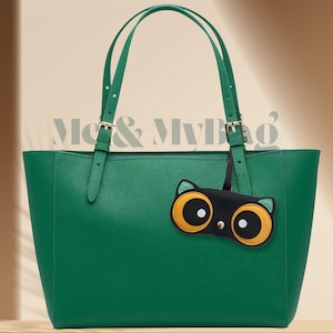 以下が含まれることがあります： 黒と黄色の猫の顔のチャームが付いた緑色のトートバッグ。バッグには2本のストラップとバックル留めがあります。