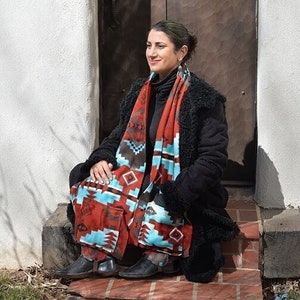 May include: A woman wearing a black jacket and a red, blue, and brown patterned scarf. The scarf is wrapped around her neck and hangs down to her waist. She is sitting on a brick step in front of a wooden door.