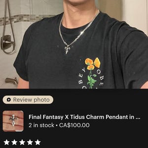 A young man wearing a black t-shirt with a charm pendant and a necklace is shown in a bathroom setting.