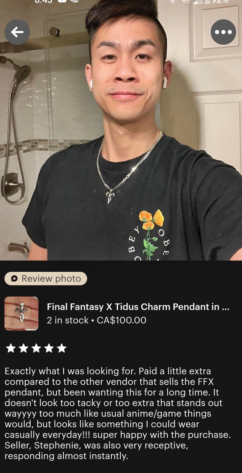 A young man wearing a black t-shirt with a charm pendant and a necklace is shown in a bathroom setting.