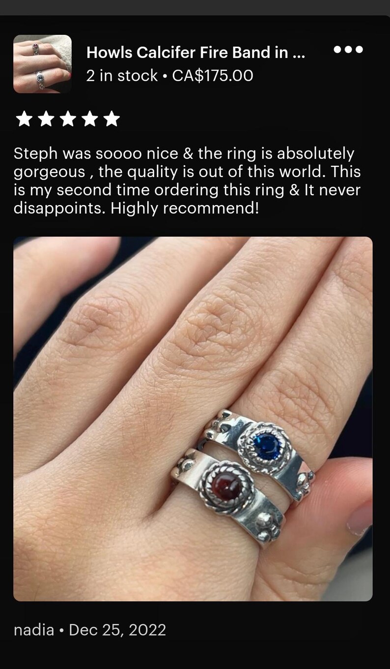 A close-up image of a person's hand wearing a silver ring with two gemstones, one blue and one red. The person is described as having a "calcifer Fire Band" in the image.