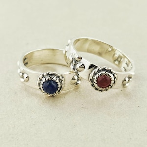 May include: Two sterling silver rings featuring a polished band with raised spherical accents. One ring is set with a round blue stone and the other with a round red stone, both encased in a twisted rope-style metal border. The interior is marked 925.