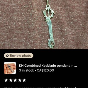 a necklace with a keyblade pendant, which is a combination of a key and a blade. The pendant is made of silver and has a unique design. The necklace is displayed on a brown fabric background.