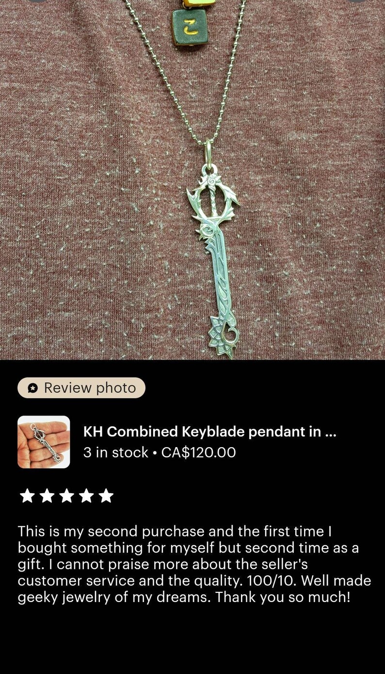 a necklace with a keyblade pendant, which is a combination of a key and a blade. The pendant is made of silver and has a unique design. The necklace is displayed on a brown fabric background.