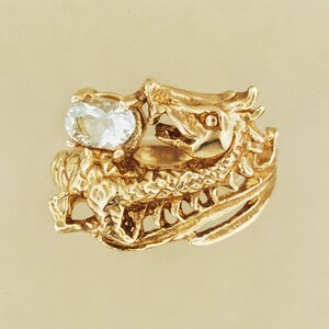 May include: A gold ring featuring a sculpted dragon design with a textured, scaled body and an open mouth. The dragon holds a single, clear, oval-cut gemstone in its claw. The metal has a polished finish, highlighting the intricate details of the creature.