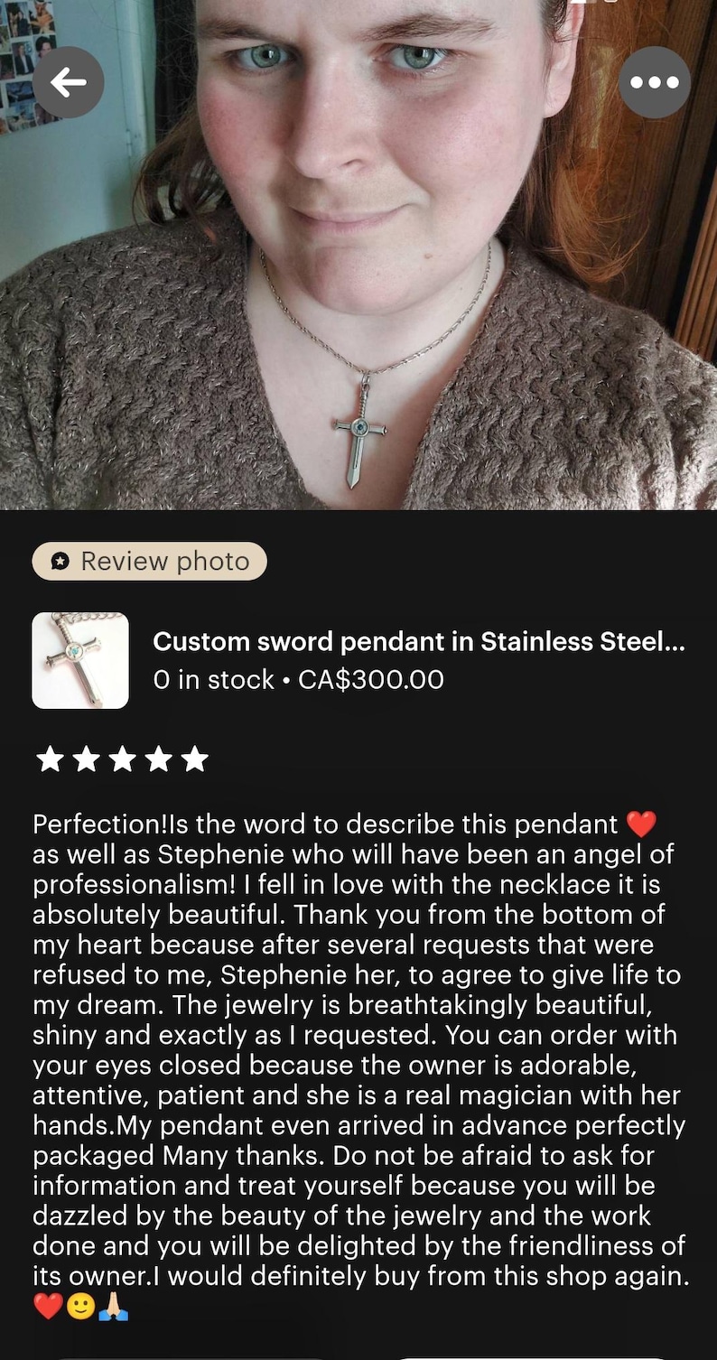 The image is a close-up of a woman's face wearing a pendant necklace with a cross pendant. The pendant is described as a custom sword pendant in stainless steel, and it is shown in stock at $300.00. The image also includes a review rating of 5 stars, indicating that the necklace is well-received by customers.