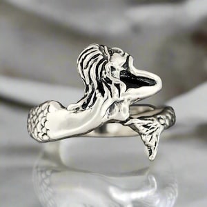 May include: A silver-toned metal ring featuring a sculpted mermaid design. The band wraps around the finger, with the mermaid's head and torso forming one end and the detailed, textured fish tail forming the other end of the open-style ring.