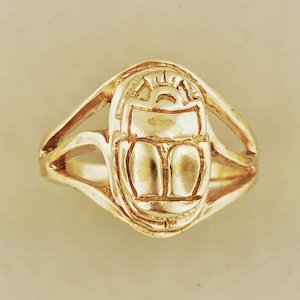 May include: A gold-toned ring featuring a central scarab beetle motif. The beetle design is embossed with a semi-circular arch at the top and segmented wing covers. The band of the ring is split into two thin, curved arms that meet at the sides.