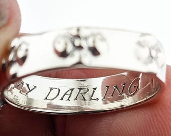 May include: A silver-toned metal ring featuring a series of embossed circular patterns on the outer band. The interior surface of the ring is clearly engraved with the text MY DARLING in a serif font, visible against the polished metallic finish.