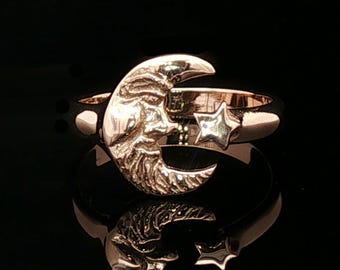 May include: A rose gold ring featuring a crescent moon with a textured, detailed surface and a small, smooth five-pointed star attached to the side of the band. The metal has a polished, reflective finish against a dark background.