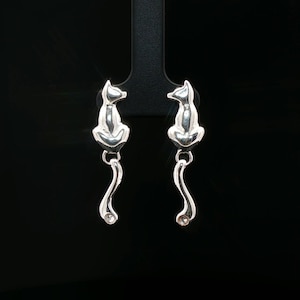 Peut inclure: Une paire de boucles d'oreilles en métal argenté brillant en forme de chats assis. Chaque boucle présente un corps félin stylisé avec une longue queue incurvée vers le bas, se terminant par une petite pierre précieuse ronde et transparente.
