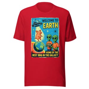 Puede incluir: Camiseta roja con un diseño gráfico de estilo retro. El diseño incluye el texto "WELCOME TO EARTH" y "HOME OF THE BEST BBQ IN THE GALAXY". El gráfico presenta un platillo volante, un cerdo, dos extraterrestres y un globo terráqueo.