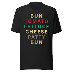 Puede incluir: Camiseta negra con las palabras "BUN, TOMATE, LECHUGA, QUESO, CARNE, PAN" en texto colorido apilado. El texto está dispuesto verticalmente, cada palabra en un color diferente, que recuerda a los ingredientes de una hamburguesa.