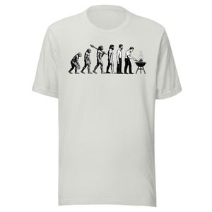 May include: A light gray t-shirt featuring a black and white graphic depicting the evolution of man, culminating in a person grilling on a barbecue. The shirt has a crew neck and short sleeves.