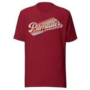 Peut inclure: T-shirt bordeaux avec le mot "Pitmaster" dans une police rétro. Le texte est crème, avec un motif à rayures multicolores en arrière-plan. Le t-shirt a des manches courtes et un col rond.