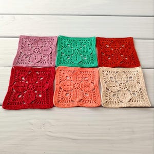 May include: Six colorful, square crochet pieces in shades of pink, green, red, coral, and beige. Each piece features a floral design in the center. The crochet squares are arranged on a light wood surface.