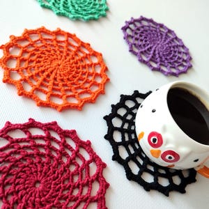 May include: A collection of colorful, crocheted doilies in orange, red, green, purple, and black. A white owl-themed mug filled with coffee rests on a black doily. The doilies are arranged on a white surface.