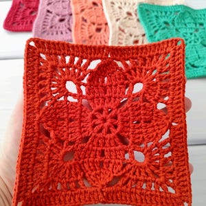 May include: A close-up of a square, crocheted doily in a vibrant orange hue, held in a hand. The doily features an intricate floral design in the center and a detailed border. Other colorful doilies in pink, peach, cream, and green are in the background.