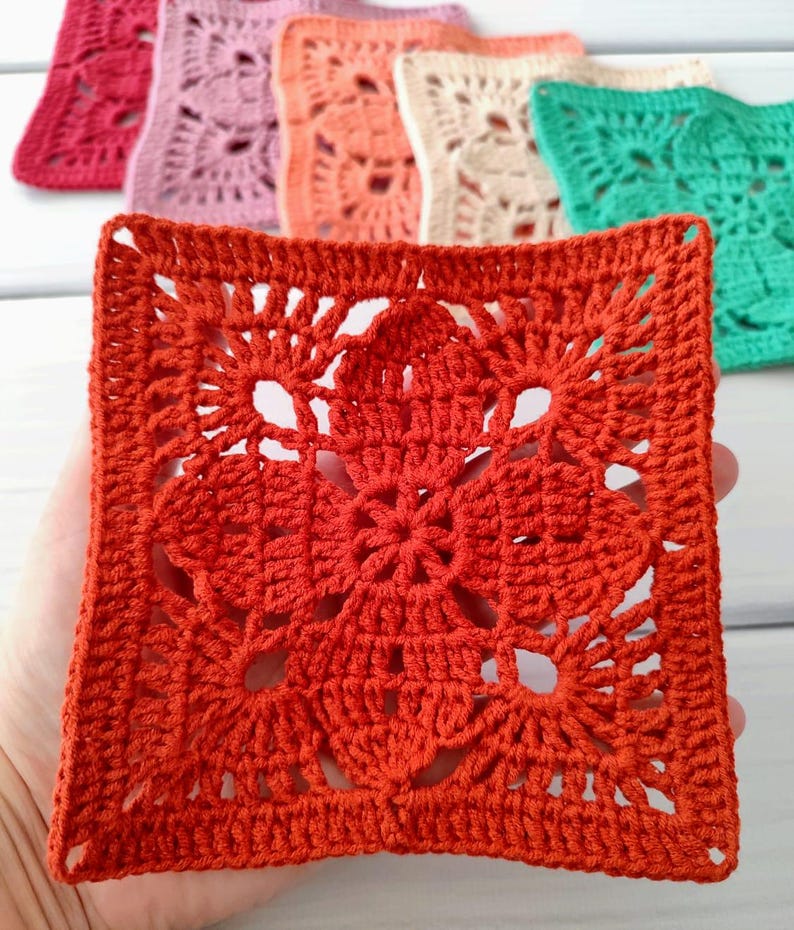 May include: A close-up of a square, crocheted doily in a vibrant orange hue, held in a hand. The doily features an intricate floral design in the center and a detailed border. Other colorful doilies in pink, peach, cream, and green are in the background.