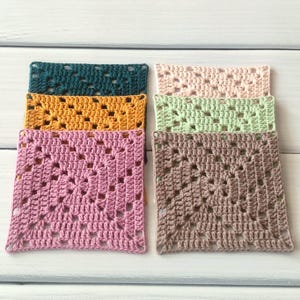 May include: A collection of six square crochet coasters arranged in two stacks. The colours include dark teal, mustard yellow, dusty pink, pale blush, light mint green, and taupe. Each coaster features a classic openwork granny square pattern.