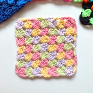 May include: A square, handmade crochet piece in a granny stitch pattern. The piece features a repeating pattern of colorful stitches in pink, yellow, green, and lavender. The edges are finished with a pink border. Other crochet pieces are visible in the background.