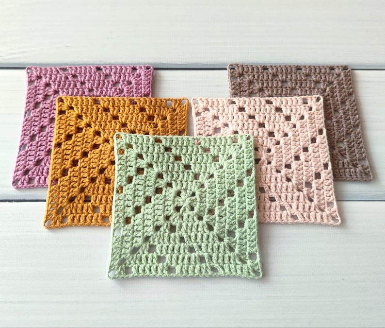 May include: Five square crochet coasters in a granny square pattern. The set includes one piece each in light pink, dusty rose, mustard yellow, sage green, and brown. Each coaster features a central flower motif with openwork stitches.