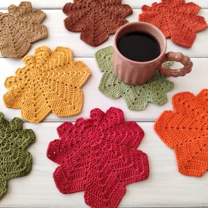 May include: A collection of handmade, leaf-shaped crochet coasters in various colors, including brown, yellow, green, orange, and red. A pink mug filled with coffee rests on a green coaster. The coasters are arranged on a white wooden surface.