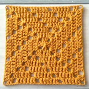 May include: A square crochet granny square in a solid mustard yellow colour. The textile features a repeating pattern of clusters of double crochet stitches separated by open chain spaces, creating a geometric, openwork grid design throughout.