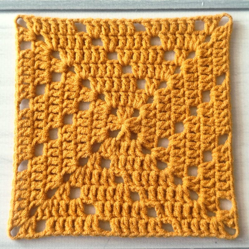 May include: A square crochet granny square in a solid mustard yellow colour. The textile features a repeating pattern of clusters of double crochet stitches separated by open chain spaces, creating a geometric, openwork grid design throughout.