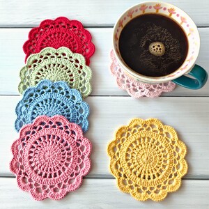 May include: A collection of colorful, handmade crochet coasters in red, green, blue, pink, and yellow. A mug of coffee sits on a pink coaster. The coasters are round with a scalloped edge and intricate openwork design.