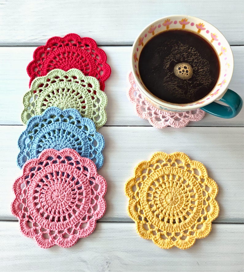 May include: A collection of colorful, handmade crochet coasters in red, green, blue, pink, and yellow. A mug of coffee sits on a pink coaster. The coasters are round with a scalloped edge and intricate openwork design.
