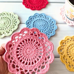 May include: A collection of handmade crochet coasters in various colors, including red, green, blue, pink, and yellow. The coasters have a circular design with a scalloped edge. A coffee cup rests on a pink coaster.