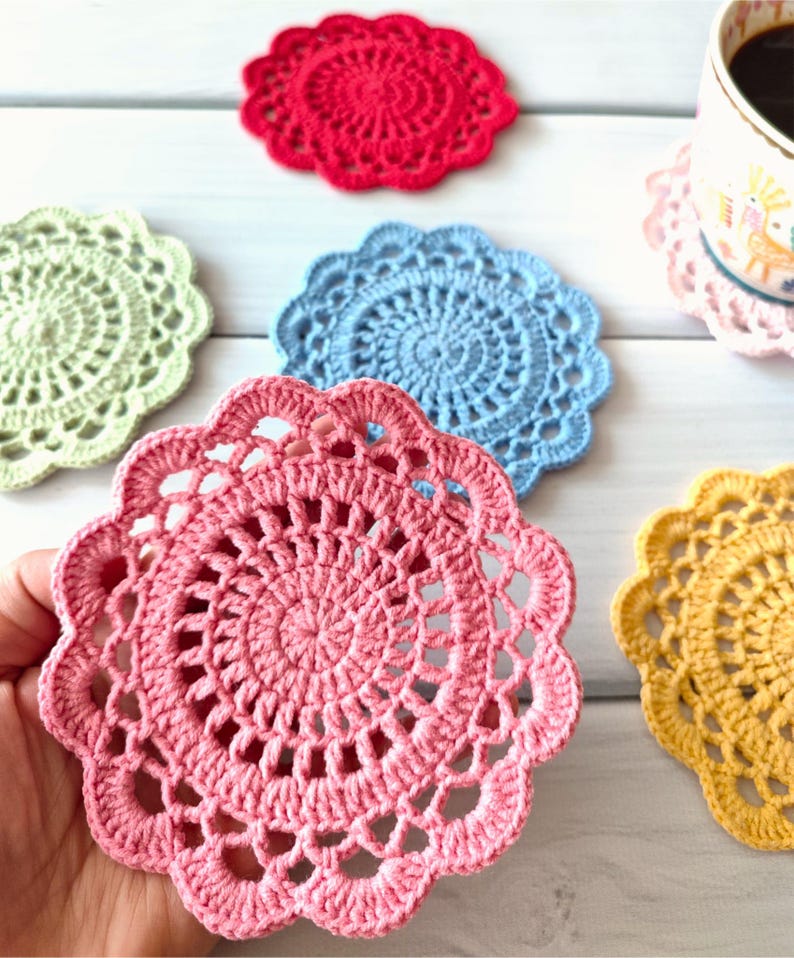 May include: A collection of handmade crochet coasters in various colors, including red, green, blue, pink, and yellow. The coasters have a circular design with a scalloped edge. A coffee cup rests on a pink coaster.