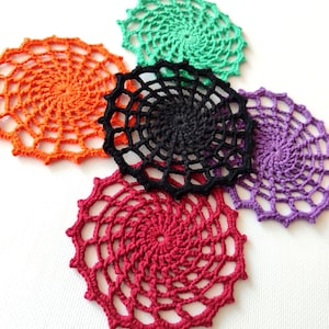 Pode incluir: Uma coleção de seis toalhinhas de crochê feitas à mão em várias cores: laranja, verde, preto, roxo e vermelho. Cada toalhinha apresenta um design em espiral com uma textura aberta e rendada. As toalhinhas estão dispostas sobre uma superfície branca.