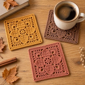 Peut inclure: Trois dessous de verre carrés au crochet en jaune moutarde, marron et orange terre cuite, chacun présentant un motif ajouré détaillé inspiré des fleurs. Une tasse en céramique remplie de café noir repose sur le dessous de verre marron.