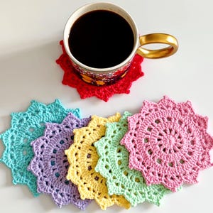 May include: A set of six round, crocheted coasters in red, turquoise, lavender, yellow, light green, and pink. Each coaster features a scalloped, star-like edge. A ceramic mug with a gold handle sits on the red coaster filled with black coffee.