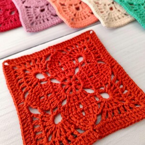 May include: Close-up of a vibrant orange crochet square with a floral design. The square is part of a set, with other squares in shades of pink, peach, beige, and green visible in the background. The crochet work is detailed and textured.