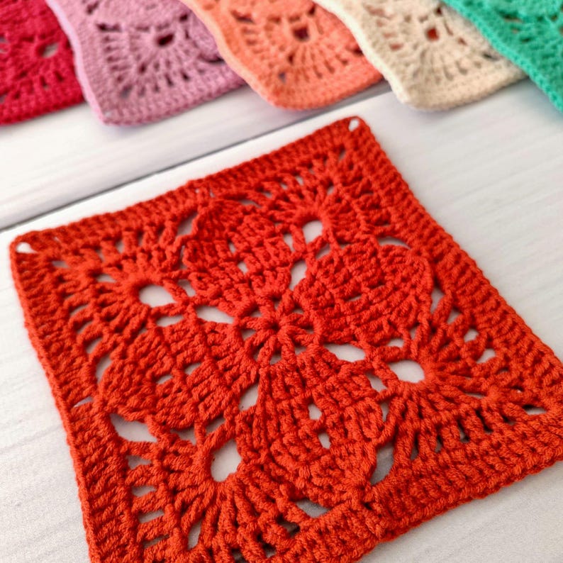 May include: Close-up of a vibrant orange crochet square with a floral design. The square is part of a set, with other squares in shades of pink, peach, beige, and green visible in the background. The crochet work is detailed and textured.