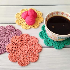 May include: A collection of handmade crochet coasters in various colors, including purple, coral, yellow, and green. A floral patterned coffee cup filled with dark coffee sits on a green coaster. Pink candies rest on a yellow coaster.