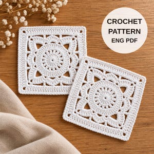 May include: Two square white crochet motifs featuring a central circular floral design with intricate petal-like stitching. A circular white label with black text reads CROCHET PATTERN ENG PDF. The motifs are arranged on a wooden surface.